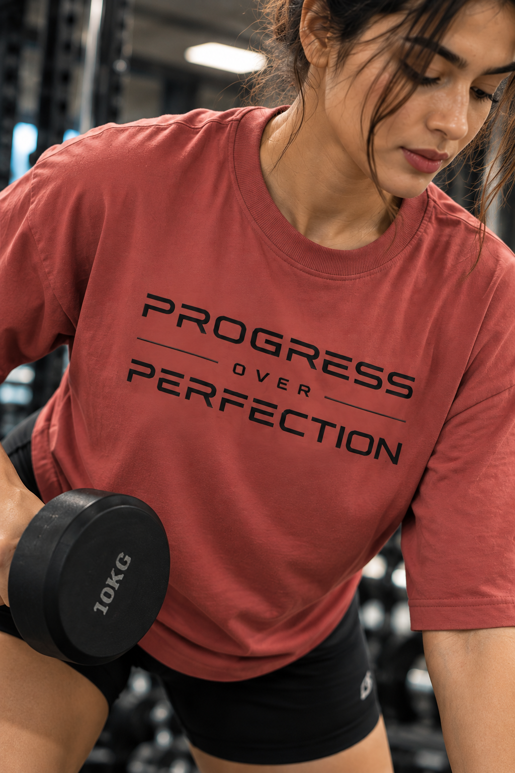 Woman performing a bench-supported dumbbell row in a muted red Progress Over Perfection T-shirt