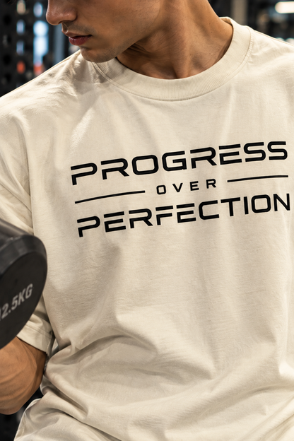 Man wearing an off-white Progress Over Perfection oversized T-shirt while curling a dumbbell in the gym