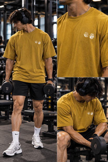 Male model wearing a mustard oversized T-shirt with a small dumbbell chest graphic, shown holding dumbbells and seated in a gym.